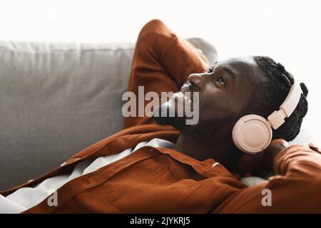 Homme noir positif dans un casque allongé sur un canapé et en écoutant de la musique, rêver et se détendre avec les mains derrière la tête Banque D'Images