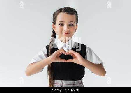 portrait de bonne écolière en chemise blanche et gilet montrant le coeur avec les mains isolées sur gris, image de stock Banque D'Images