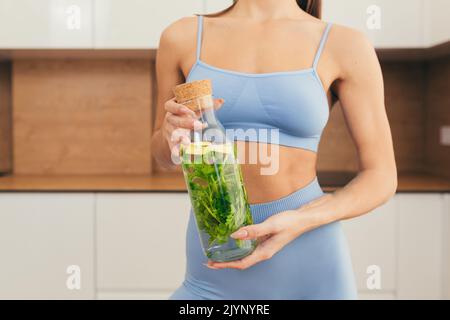 Gros plan photo partie du corps de la jeune femme de forme physique tenant et montrant une bouteille de jus frais, détox à la maison Banque D'Images