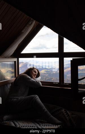 Jeune femme assise dans une maison confortable et regardant la fenêtre. Fond de montagne Banque D'Images