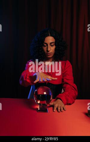 brunette fortune teller avec les yeux fermés tenant la main sur boule de cristal sur fond sombre avec drapé rouge Banque D'Images