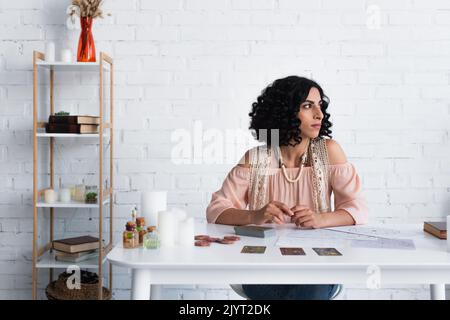 brunette fortune teller regardant loin en s'asseyant près des cartes de tarot et des cartes cosmiques Banque D'Images