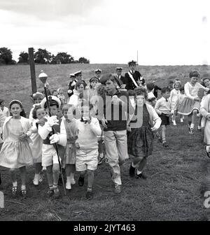 1965, historique, enfants à l'extérieur dans un champ près d'un domaine de logement prenant part au gala du North Queensferry, Fife, Écosse, Royaume-Uni, les enfants suivent un garçon habillé comme un tambour majeur tenant un mace. Banque D'Images