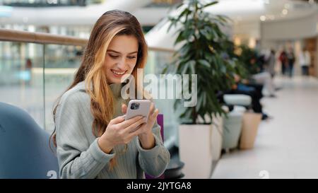 Gai heureux jeune fille shopper shopaholic femme assis dans le centre commercial avec des paquets regardant le téléphone victoires se réjouit des réductions réussi de commande Banque D'Images