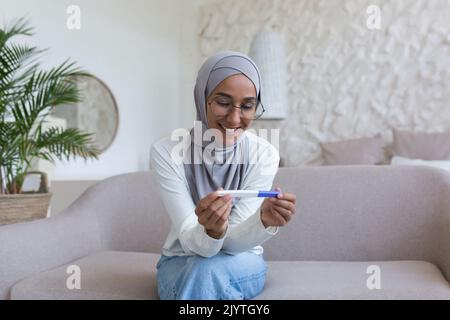 Femme musulmane heureuse et souriante dans le hijab tenant un test de grossesse à la maison, femme arabe heureuse de résultat assis sur un canapé dans le salon. Banque D'Images