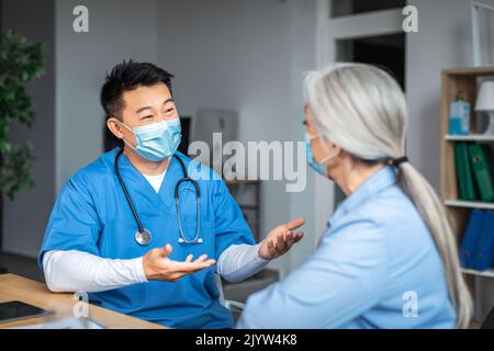 Thérapeute japonais d'âge moyen heureux dans un masque de protection parlant avec une vieille femme patiente à l'intérieur du bureau de la clinique Banque D'Images