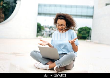 Une jeune femme afro-américaine satisfaite, aux cheveux mauriquement, avec micro-casque, travailleur de l'entreprise, consultation en ligne, négociations avec des collègues, assis à l'extérieur, utilisation d'un ordinateur portable, conversation vidéo, sourire Banque D'Images