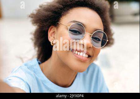 Moderne tendance joyeuse africaine fille américaine avec des cheveux bouclés, avec des lunettes bleues, blogueur vidéo, prend un selfie sur le téléphone portable, regarde la caméra frontale, enregistre le flux en ligne, sourire heureux Banque D'Images