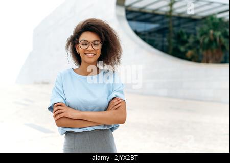 Femme d'affaires réussie. Portrait d'une femme afro-américaine élégante et confiante avec des cheveux bouclés, en chemise bleue, avec des lunettes, debout à l'extérieur avec les bras croisés, regarde l'appareil photo, sourit amicaux Banque D'Images