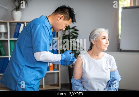 Adulte sérieux homme asiatique médecin faire l'injection à la vieille femme patiente dans l'intérieur du bureau de la clinique Banque D'Images