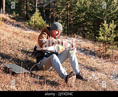 Jeune homme en vêtements d'automne buvant du thé d'un thermos tout en étant assis au bord de la forêt en soirée au soleil se reposant en randonnée et en appréciant la nature Banque D'Images