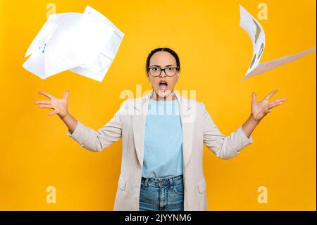 Une dame d'affaires brésilienne ou latinos irrité avec des lunettes, scanne des documents financiers en colère, regarde la caméra, se tient sur un fond orange isolé, expression faciale émotionnelle Banque D'Images