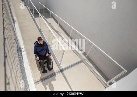 Homme en fauteuil roulant, approchant le bâtiment le long d'une rampe accessible pour les personnes handicapées Banque D'Images