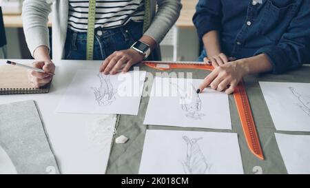 Gros plan des mains des femmes designers qui choisissent des croquis pour la nouvelle collection de vêtements. Des dessins de vêtements tendance sont sur la table avec le tissu et les accessoires de couture. Banque D'Images