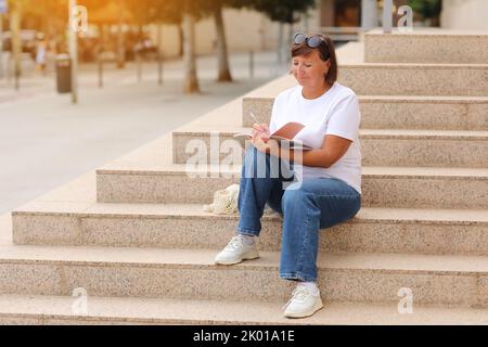 La femme mature travaille à distance, l'enseignement supérieur pour les adultes, la femme décontractée écrit et note dans un carnet assis sur les escaliers à l'extérieur, freelance.C Banque D'Images