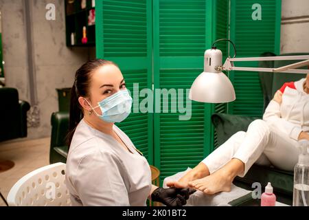 Jeune podiatre femelle avec un masque protecteur faisant de la chiropodie et regardant la caméra dans sa clinique de podiatrie Banque D'Images