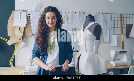 Portrait à la taille haute de la belle jeune femme tailleur debout sur le lieu de travail près d'un mannequin vêtu et regardant l'appareil photo. Les vêtements, les croquis et les machines à coudre pour femmes sont visibles. Banque D'Images