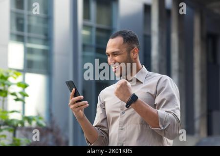 Homme d'affaires afro-américain à l'extérieur du bâtiment de bureau utilisant le téléphone, souriant et heureux tenant main vers le haut le geste de triomphe, jeune entrepreneur célébrant la victoire lisant de bonnes nouvelles d'investissement en ligne. Banque D'Images