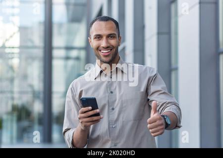 Jeune homme d'affaires afro-américain à l'extérieur du bâtiment de bureau souriant et regardant la caméra, pouces vers le haut, programmeur utilisant un smartphone pour tester des applications. Banque D'Images