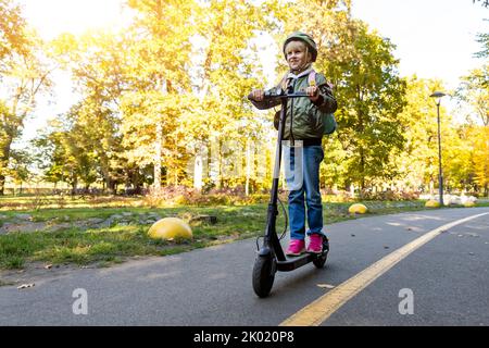 Profil vue portrait de mignon blond petite école caucasienne fille port casque profiter d'avoir plaisir à l'équitation électrique scooter ville parc de rue à l'extérieur sur Banque D'Images