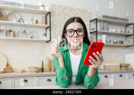 Jeune femme nerveuse dans des lunettes tenant un téléphone rouge et regardant avec impatience un appel ou un message important. À la maison dans la cuisine, regardant l'appareil photo, inquiet. Banque D'Images