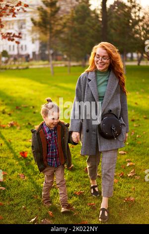 Femme souriante et petit garçon marchant sur une pelouse en herbe verte dans le parc de la ville le chaud jour d'automne. Jeune maman va avec son fils, ils ont du plaisir et de tenir Banque D'Images