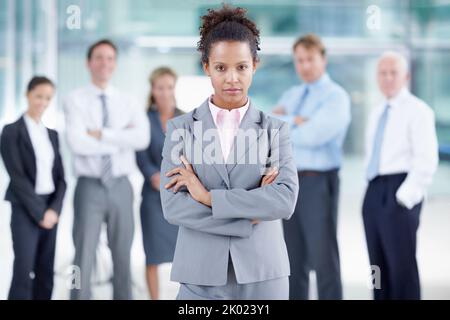 Prendre sa carrière au sérieux. Sérieuse jeune femme d'affaires africaine debout avec ses collègues derrière elle - portrait. Banque D'Images