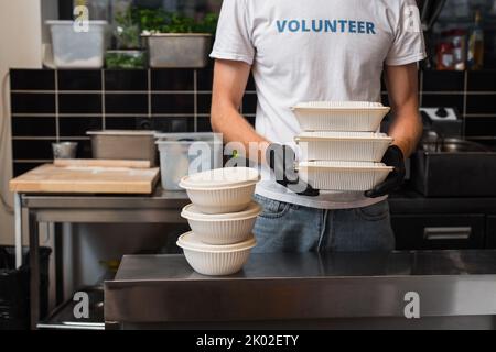 vue rognée de l'homme dans un t-shirt avec lettrage volontaire tenant des récipients en plastique dans la cuisine, image de stock Banque D'Images