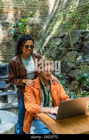 bonne femme afro-américaine touchant les épaules de l'homme à tête rouge en utilisant un ordinateur portable en terrasse extérieure, image de stock Banque D'Images