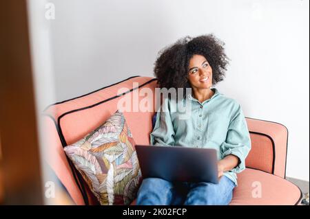 Femme afro-américaine optimiste en tenue décontractée utilisant un ordinateur portable tout en étant assise sur le canapé. Jeunes étudiantes regardant des webinaires, des cours éducatifs, apprenant à distance, tapant des e-mails Banque D'Images