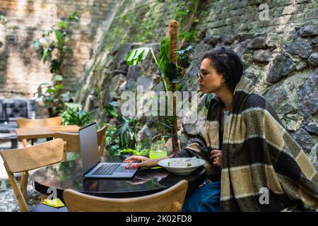 travailleur indépendant afro-américain couvert dans une couverture avec ordinateur portable et déjeuner dans un café sur la terrasse, image de stock Banque D'Images