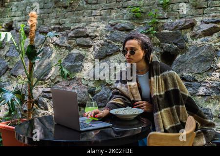 travailleur indépendant afro-américain dans des lunettes de soleil couvertes dans une couverture en utilisant un ordinateur portable et ayant déjeuner dans un café sur la terrasse, image de stock Banque D'Images