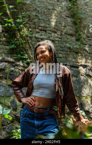 bonne femme afro-américaine dans une veste en cuir marron debout avec la main sur la hanche, image de stock Banque D'Images