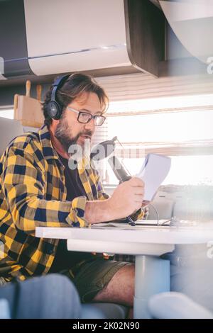 Homme adulte moderne travaillant avec un ordinateur portable et un microphone dans une camionnette de camping dans le mode de vie de travail nomade numérique. Technologie et voyages. Concept de bureau de liberté alternatif avec nouvelle activité et personnes libres Banque D'Images