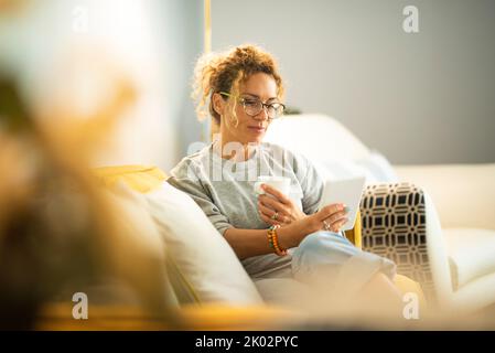 Jeune femme caucasienne adulte détendue a lu un livre sur un lecteur d'appareil moderne confortablement assis sur le canapé à la maison. Portrait de jolies femmes portant des lunettes et utilisant la connexion Banque D'Images