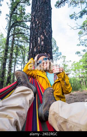 Liberté et aventure voyage style de vie pour un couple adulte dans la forêt de bois de la nature - femme debout regarder le fond du parc extérieur et de boire - homme couché sur hamac et de repos Banque D'Images