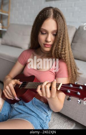 jeune fille ciblée dans des vêtements décontractés jouant ukulele dans la salle de séjour moderne, image de stock Banque D'Images