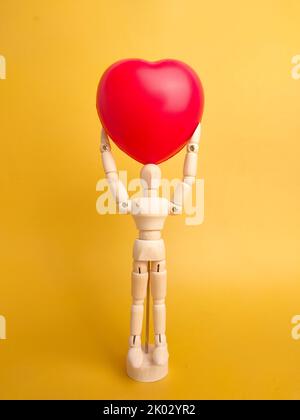 Un mannequin en bois tenant un coeur rouge sur un fond jaune. Banque D'Images
