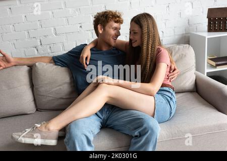 bonne adolescente en train de mettre des jambes sur un ami souriant tout en étant assis sur un canapé, image de stock Banque D'Images