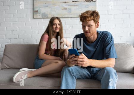 jeune homme à tête rouge utilisant un smartphone tout en s'asseyant près d'une petite amie souriante sur un canapé, image de stock Banque D'Images