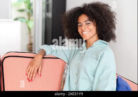 Portrait d'une jeune femme souriante dans une tenue décontractée assis sur le canapé confortable dans une posture détendue et regarde l'appareil photo, femme calme et insouciante multiraciale dans un appartement moderne Banque D'Images