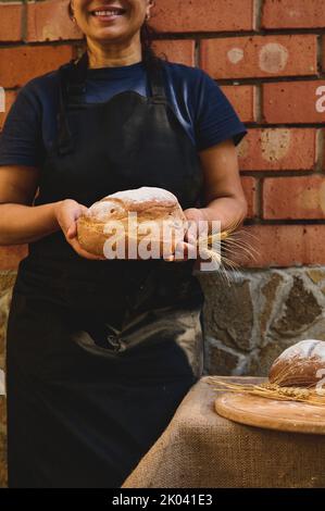 Femme charmante, boulanger en tablier de chef noir et casquette blanche tenant un pain de pain de levain brun de grain entier fait maison Banque D'Images