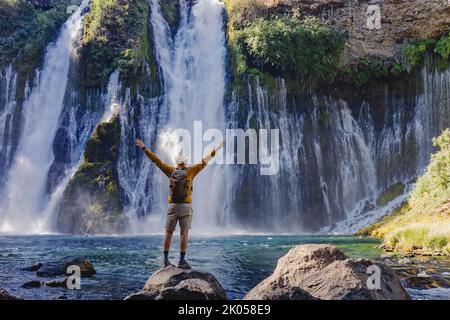Homme voyageur debout avec ses mains levées avec une vue sur la belle cascade, vue de l'arrière Banque D'Images