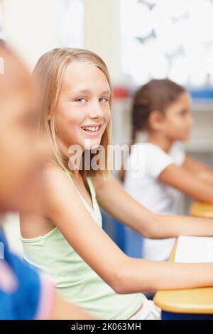 Je me sens confiant dans ma classe. Jeune fille blonde regardant l'appareil photo tout en étant assise à son bureau en classe - Copyspace. Banque D'Images