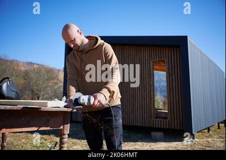 Homme chauve de coupe de métal, à l'aide d'une scie circulaire tout en construisant une maison de cadre en bois dans le style scandinave le jour ensoleillé avec fond bleu ciel. Construction de la maison de grange concept. Banque D'Images