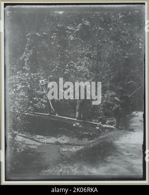 Passerelle de traversée de groupe avec caméras, entre 1885 et 1900. Photographie montrant deux hommes transportant des appareils photo et des trépieds aidant une femme à traverser une passerelle en rondins. Banque D'Images