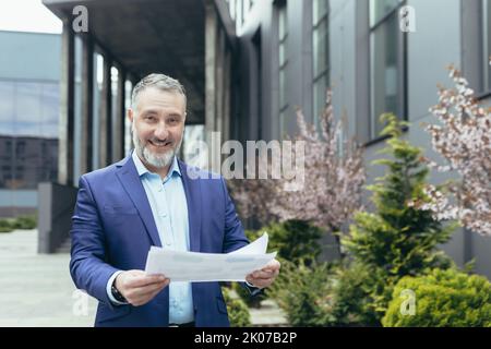 Agent immobilier. Un beau homme âgé et une barbe se tiennent dans la rue près des maisons, du bureau, tenant des documents entre ses mains, satisfait d'une affaire réussie. Regarde la caméra, sourit. Banque D'Images