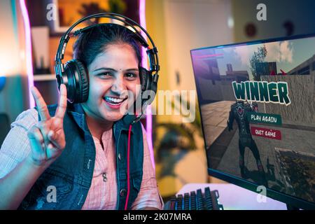 Joueur souriant avec casque célébrant le jeu gagnant en montrant le signe de victoire tout en jouant à un jeu vidéo en direct sur ordinateur à la maison - concept de championnat Banque D'Images