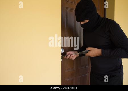 Image d'un voleur tout vêtu de noir avec une balaclava qui tente de forcer la serrure d'une porte de maison avec un gros tournevis. Augmentation constante à la maison Banque D'Images
