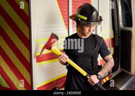 Photo d'un pompier portant un casque avec une hache contre un moteur d'incendie. Banque D'Images